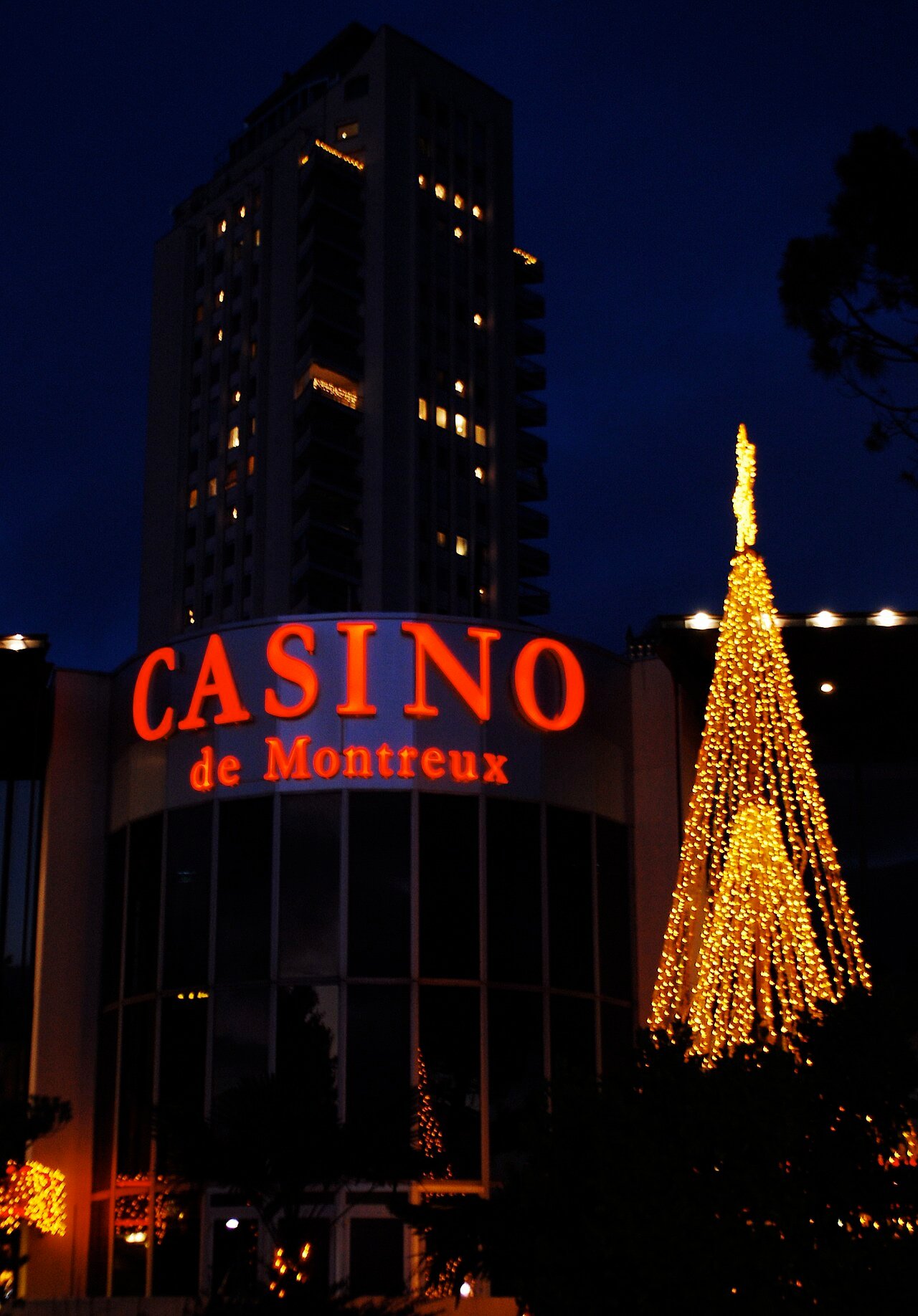The red neon sign of the Casino de Montreux at night, with a lit Christmas tree beside the entrance