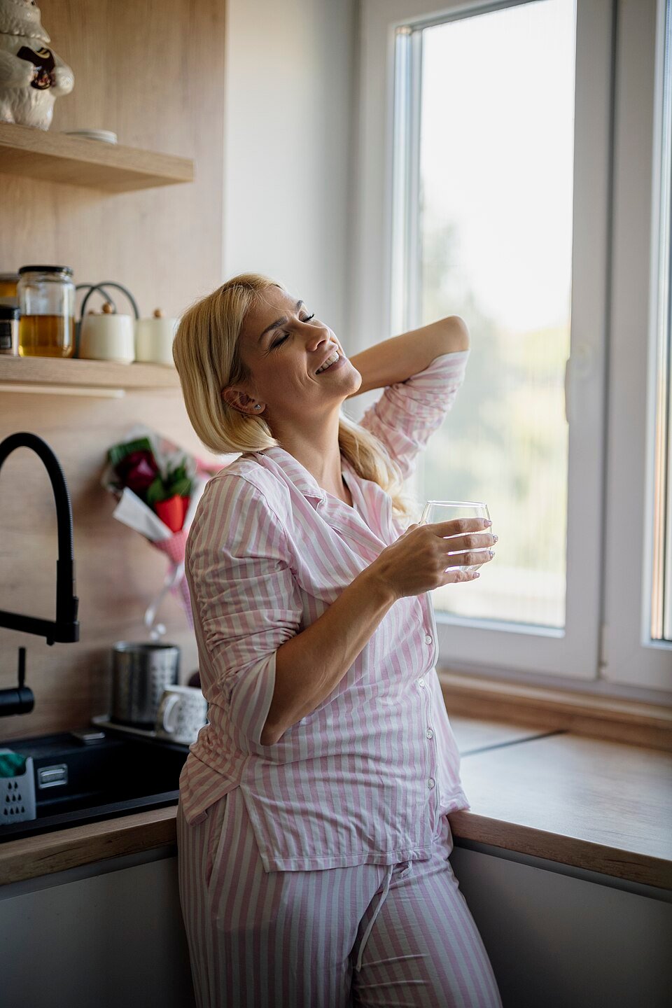 A woman in striped pyjamas stands by a sunny kitchen window in the morning, holding a glass of water