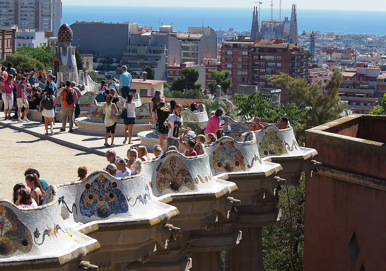 The main terrace of Park Güell in Barcelona with its curving mosaic bench and visitors, and the city, the Sagrada Família and the sea in the distance