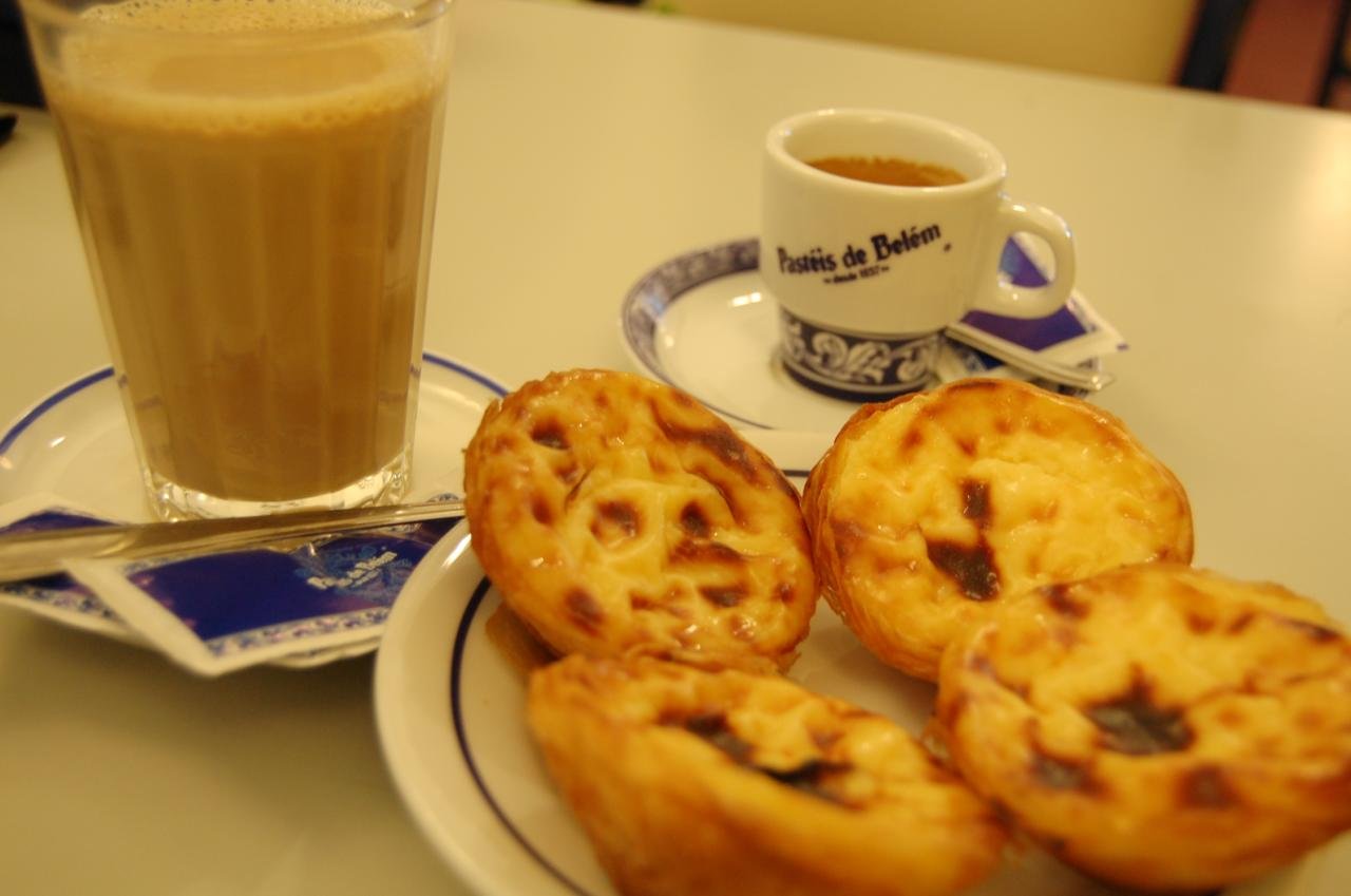 Pastéis de Belém custard tarts on a plate with a coffee cup bearing the Pastéis de Belém name and a glass of milky coffee
