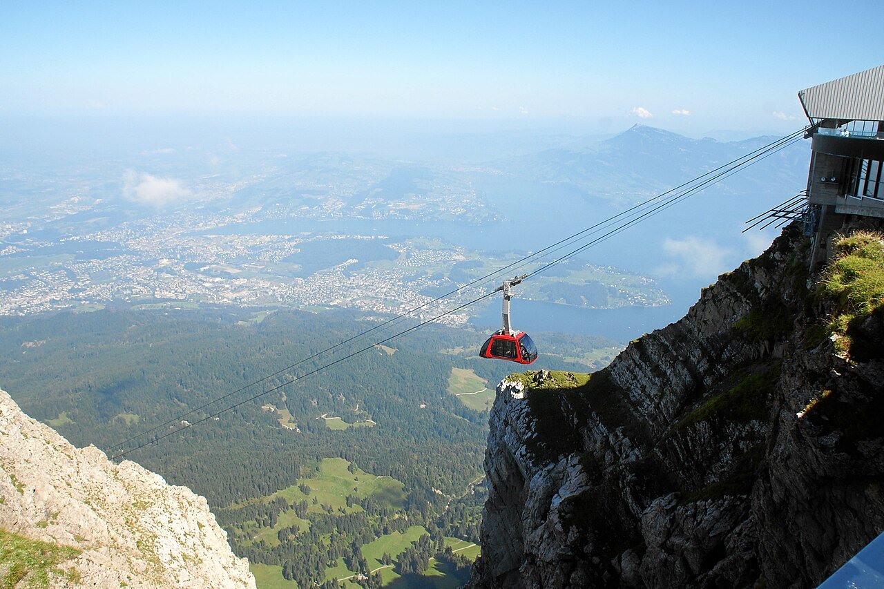 A red cable car hanging over a steep drop below the rocks of Mount Pilatus, with the Lucerne area and lakes far below