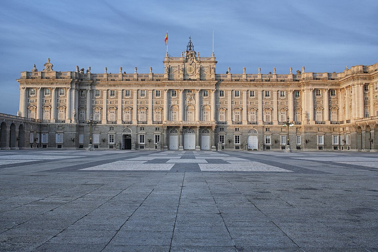 The south façade of the Royal Palace of Madrid seen across the wide paved Plaza de la Armería under a blue sky