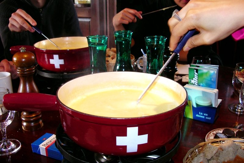 A red pot of melted cheese fondue with a Swiss cross on its side, and diners dipping bread on long forks