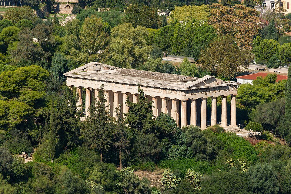 The Temple of Hephaestus, a Doric temple with its columns intact, among green trees in the Ancient Agora of Athens, seen from the Acropolis