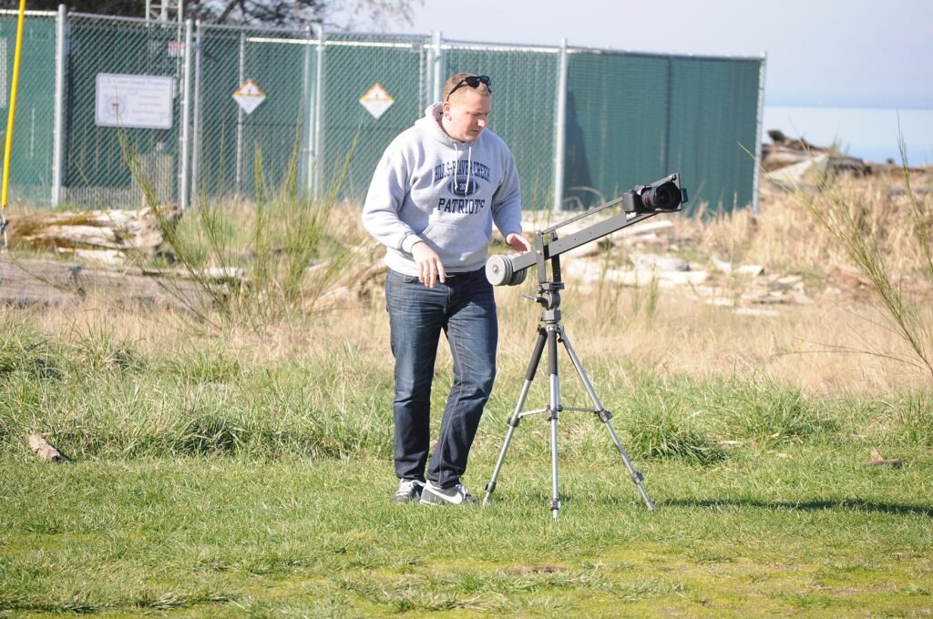 A videographer on a grassy shore adjusts a camera mounted on a tripod and a small crane arm