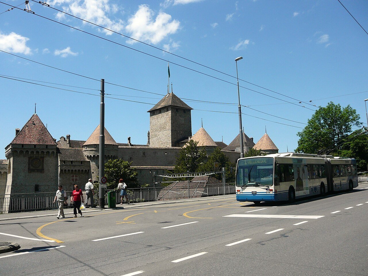 A blue and white articulated trolleybus on the lakeside road passing the towers of Chillon Castle