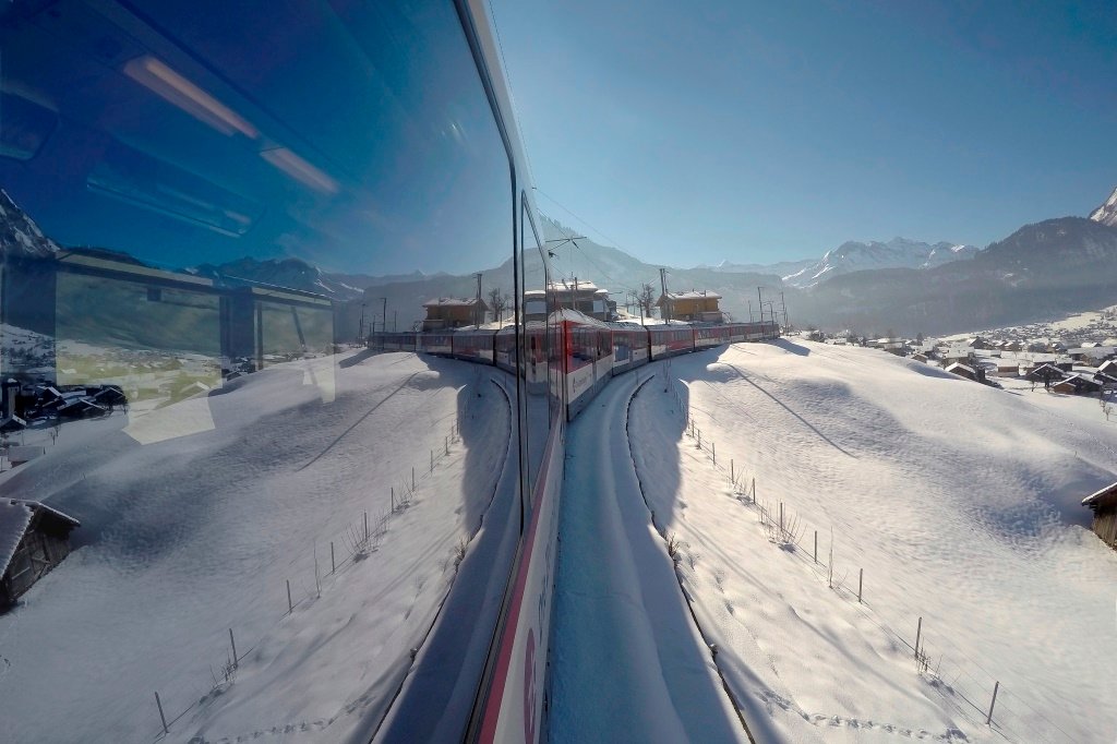 A red Zentralbahn train running through deep snow beside Lake Lungern on the Brünig line, mountains behind