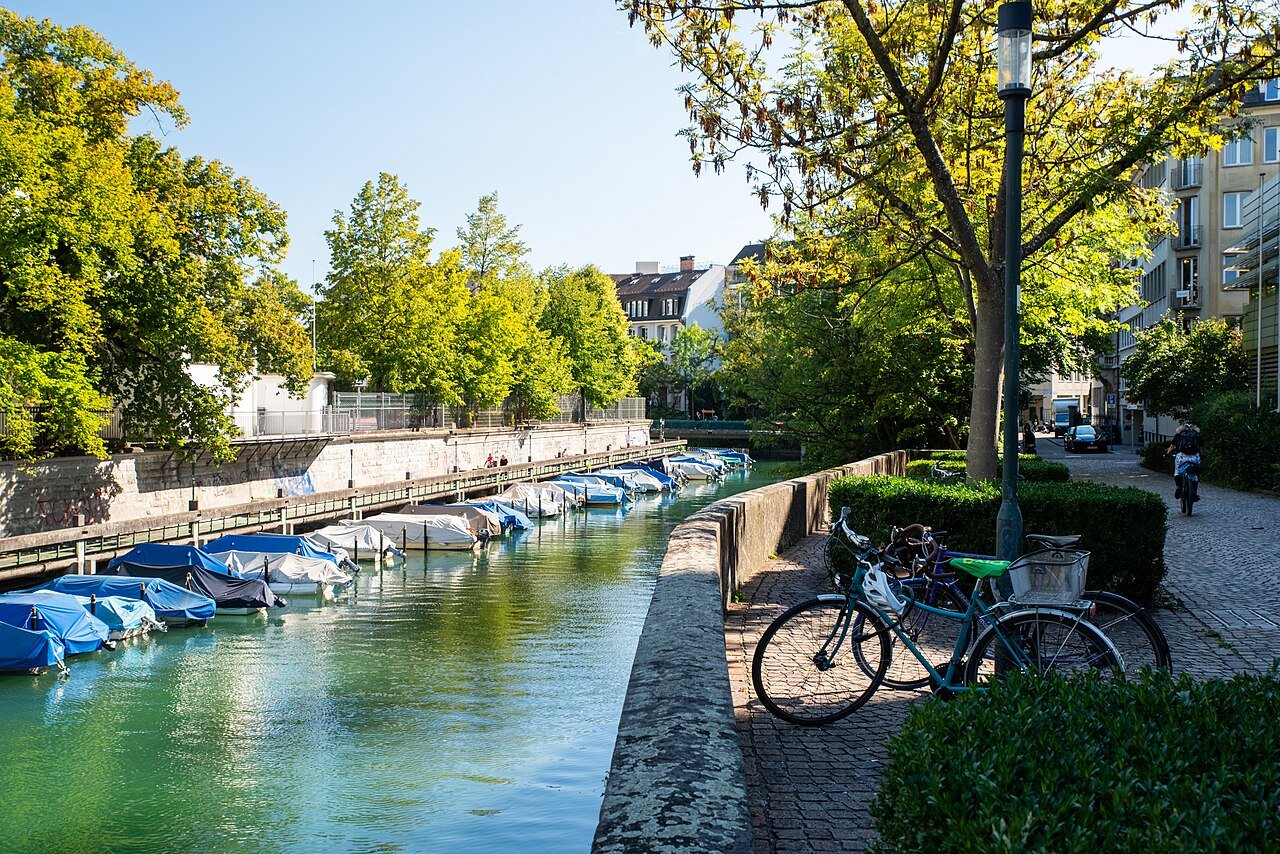 The Schanzengraben canal in Zurich, green with tree reflections, with small boats under covers moored along one side